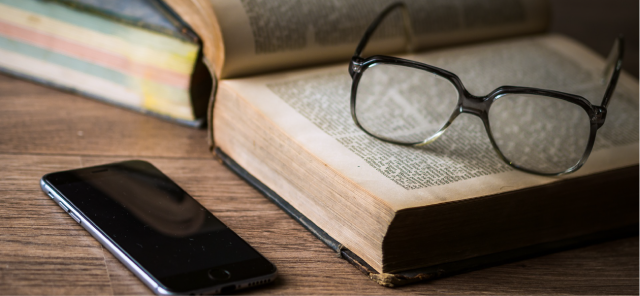 A pair of glasses resting on an open book on top of a desk, next to a smartphone