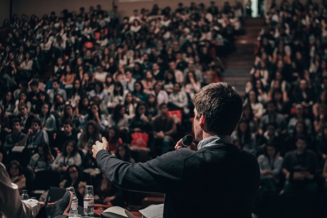 A man on a stage with a microphone, addressing many people in the audience